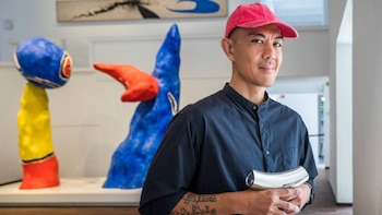 Un hombre asiático con gorra roja y camisa oscura sostiene un objeto plateado curvado, con esculturas abstractas de colores azul, rojo y amarillo en el fondo de una galería