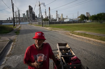 Un hombre con gorra roja de 'Trump' y camisa roja camina por una calle, empujando un carrito. Al fondo, una planta industrial con varias chimeneas humeantes