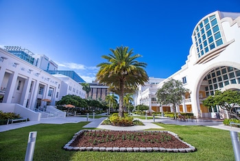 Vista exterior de un campus con edificios blancos, columnas, arcos y grandes ventanas, además de palmeras, césped y jardines bajo un cielo azul