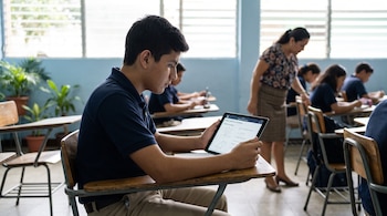 Un estudiante sentado en un escritorio de madera observa una tableta digital con ejercicios matemáticos dentro de un aula.