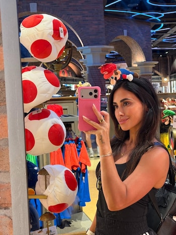 Antonela Roccuzzo, vestida de negro, se mira en un espejo mientras toma una selfie con un celular rosa. Lleva una diadema de Mario y hay peluches de Toad