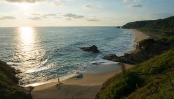 Vista panorámica de una playa arenosa con una persona, el océano con olas, rocas, y una costa con colinas cubiertas de vegetación al atardecer