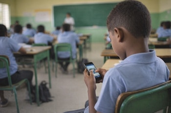 Plano trasero de un niño con uniforme escolar azul, viendo su teléfono móvil en un aula con otros estudiantes, pupitres y una pizarra verde.