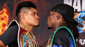 Dos boxeadores vistos de perfil se miran de cerca mientras llevan cinturones de campeonato sobre sus hombros.