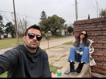 Un hombre de lentes de sol y sudadera negra toma una selfie con una mujer de denim, lentes de sol y botas, sentada junto a una pared de ladrillo. Fondo urbano con árboles y cielo nublado