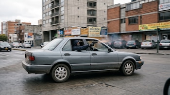 Un automóvil gris en movimiento con un hombre encapuchado que apunta una pistola desde la ventanilla hacia la calle.