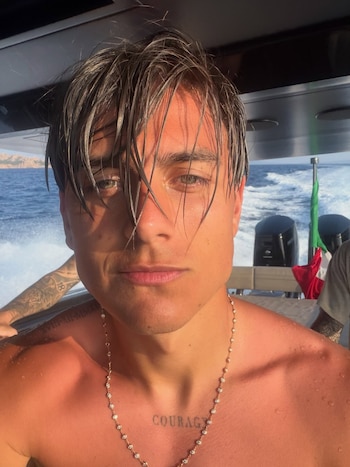 Primer plano de Paulo Dybala con el pelo mojado, torso desnudo, collar y tatuajes. Detrás, el mar con estela, un barco con motores y una bandera italiana