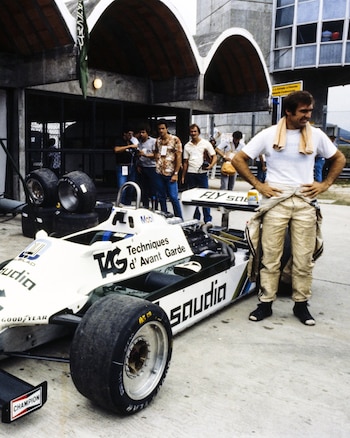 El piloto Carlos Reutemann, con camiseta blanca y toalla naranja al cuello, de pie junto a un monoplaza blanco de F1 en un box, con neumáticos detrás
