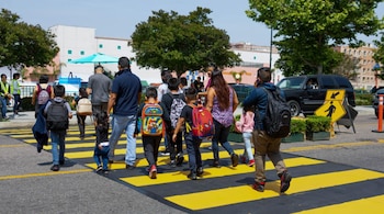 Un grupo de adultos y niños con mochilas cruza un paso de cebra amarillo y negro en una calle, con edificios y árboles verdes bajo un cielo azul claro