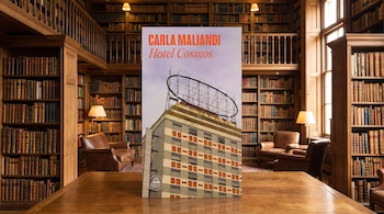Un libro titulado Hotel Cosmos de Carla Maliandi sobre una mesa de madera en una biblioteca con estanterías de libros y sillones de cuero.