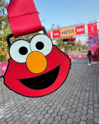 Una medalla con el rostro de Elmo en primer plano y una estructura con la palabra META al fondo