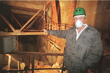 Un hombre con casco verde y mascarilla blanca señala hacia un espacio oscuro y dañado con vigas metálicas, andamios y escombros, incluyendo madera