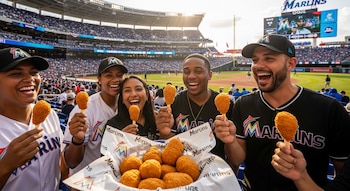 Cinco personas sonrientes sostienen patitas de pollo en forma de muslo en un estadio de béisbol con gradas llenas y el campo de juego de los Miami Marlins al fondo.