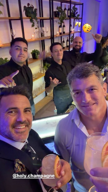 Grupo de cinco hombres en un bar con iluminación púrpura. Dos en primer plano sonríen y sostienen bebidas con rodajas de pomelo. Tres hombres en la barra detrás