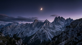 Paisaje montañoso con picos rocosos y vegetación oscura, cielo nocturno con nubes dispersas, estrellas y un sol parcialmente eclipsado de forma creciente.