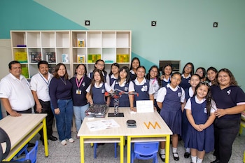 Grupo de estudiantes y adultos posan en un aula moderna con estantes llenos de kits de robótica. Sobre una mesa, un modelo de grúa y componentes tecnológicos