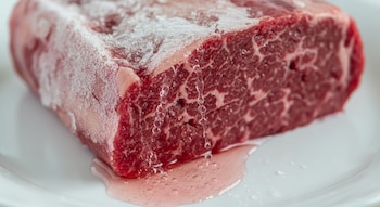 Un trozo grande de carne roja cruda, parcialmente cubierto de hielo y escarcha, se descongela sobre un plato blanco, con gotas de líquido cayendo.