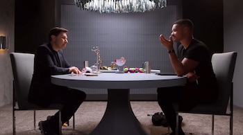 Dos hombres sentados frente a frente en una mesa redonda gris; uno viste traje negro y el otro camiseta negra, con modelos científicos sobre la mesa