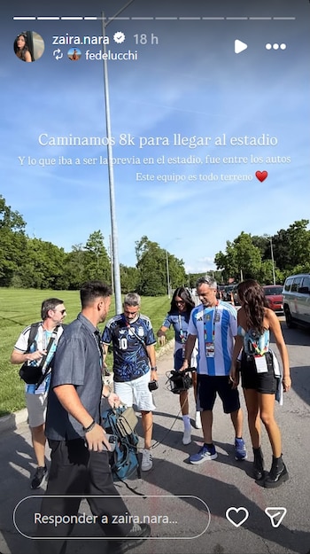 Zaira Nara y un grupo de personas, varias con camisetas de Argentina, caminan por un sendero asfaltado junto a un campo verde con árboles, bajo un cielo claro