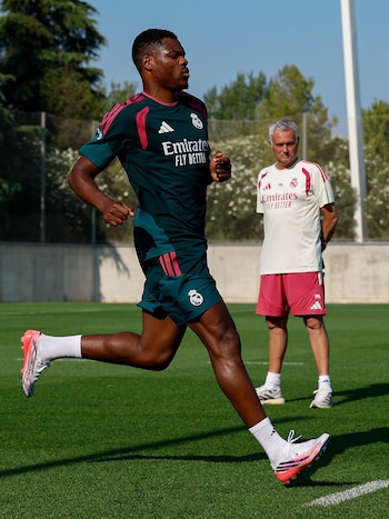 Un jugador del Real Madrid con uniforme verde corre en un campo de césped; un hombre con camiseta blanca y pantalón rojo del Real Madrid lo observa de pie