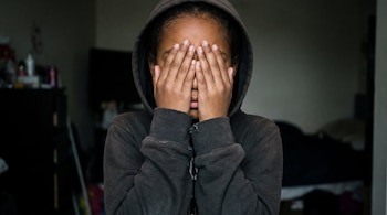 Una niña de piel morena viste una sudadera oscura con capucha y se tapa la cara con las dos manos. El fondo de la imagen está oscuro.
