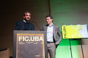 Dos hombres en un escenario detrás de un atril con la inscripción FIC.UBA y junto a una pantalla digital
