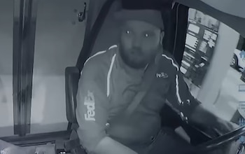 Un hombre barbudo con gorra y uniforme de FedEx está sentado al volante de un vehículo, visto desde una cámara interior. La imagen es monocromática