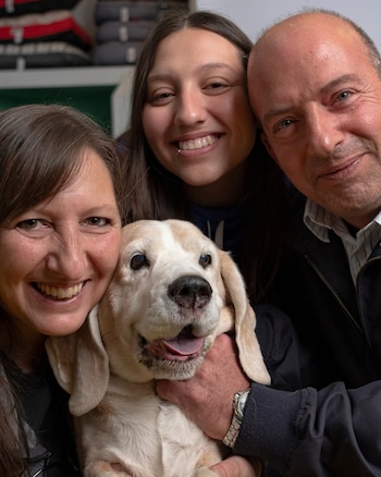 Un hombre, una mujer y una joven sonríen junto a un perro de pelaje blanco y castaño en un primer plano