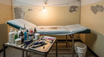 Interior de un consultorio improvisado con paredes descascaradas, camilla médica, una mesa con botellas, envases y herramientas de estética, y un cubo.