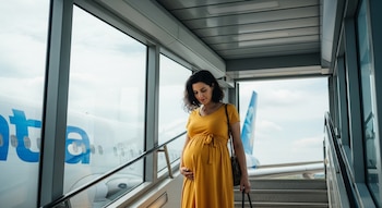 Una mujer embarazada con un vestido amarillo sube las escaleras de una pasarela de embarque. Se observa un avión a través de los ventanales con nubes en el cielo.