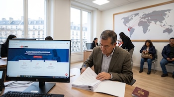 Un hombre sentado en un escritorio escribe en papeles, frente a un monitor de computadora, con un pasaporte al lado y un mapa mundial en la pared