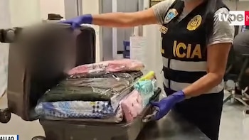 Persona con chaleco policial y guantes azules inspecciona una maleta abierta que contiene prendas de vestir envueltas en plástico