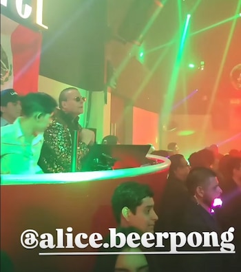 Hombre con chaqueta de lentejuelas y gafas de sol en una cabina de DJ, con luces láser verdes y rojas iluminando el fondo oscuro de una discoteca