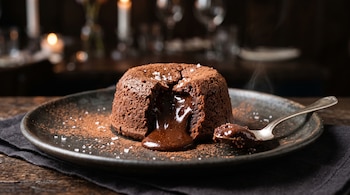 Volcán de chocolate abierto con centro líquido, espolvoreado con cacao y sal sobre un plato oscuro junto a una cuchara.