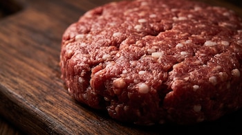 Medallón redondo de carne picada cruda con pequeños fragmentos de grasa blanca sobre una tabla de madera oscura.