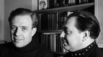 Dos hombres en blanco y negro, uno mirado al frente y otro de perfil, situados delante de una estantería con libros.