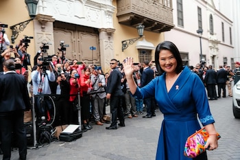 Mujer con vestido azul y bolso colorido saluda a la cámara, rodeada por una multitud de personas, periodistas con cámaras y personal de seguridad