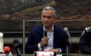 Un hombre maduro con saco azul habla sosteniendo un micrófono en una mesa junto a otros micrófonos de medios de comunicación
