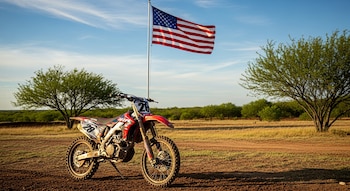 Moto de motocross roja y blanca, número 28, en camino de tierra. Bandera de Estados Unidos en asta, árboles verdes y cielo azul.