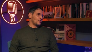 Hombre de tez clara y barba de perfil, usando camiseta oscura y micrófono de solapa. Detrás hay un letrero de neón con forma de micrófono y una estantería con libros