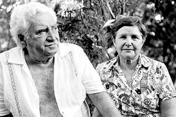Foto en blanco y negro de un hombre y una mujer sentados, el hombre con camisa abierta y la mujer con camisa estampada, con vegetación de fondo