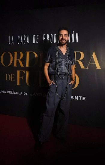 Hombre con barba y vestimenta oscura de tela satinada posa ante un fondo negro con texto dorado y blanco. Lleva un collar largo de cuentas negras
