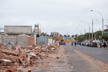 Escombros de ladrillo acumulados en la acera de una avenida donde hay maquinaria pesada, personal con uniforme y vehículos
