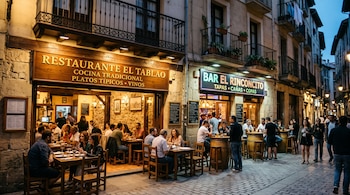 Fachadas iluminadas de un restaurante y un bar con clientes sentados en mesas y barriles de madera en una calle adoquinada.