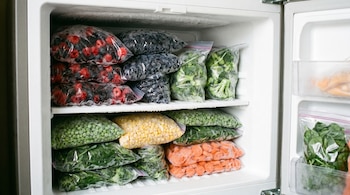 Interior de un congelador con bolsas de frutas y verduras congeladas apiladas en estantes. Se ven bayas, arándanos, brócoli, guisantes, maíz y zanahorias.