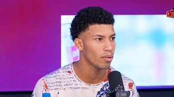 Hombre de cabello rizado con camiseta estampada frente a un micrófono en un estudio con fondo de colores morado y azul.