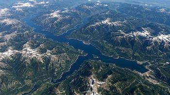 Vista aérea de un embalse azul rodeado de montañas verdes con picos nevados y una presa en el extremo inferior derecho.