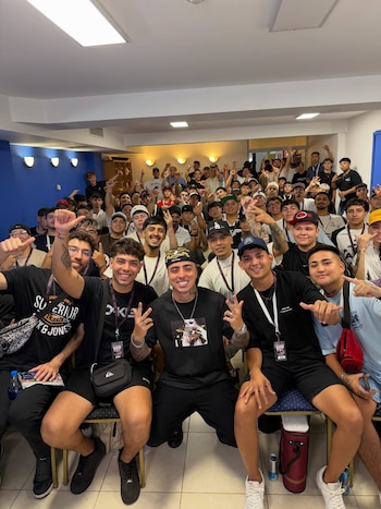 Un grupo numeroso de hombres jóvenes sonríe y posa para la cámara. Muchos llevan gorras y algunos hacen señales de mano. Fondo azul y blanco