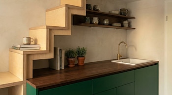 Vista de una cocina compacta bajo una escalera de madera clara, con encimera de madera oscura, fregadero blanco, grifo dorado y gabinetes verdes.