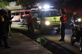 Una ambulancia con luces de emergencia, personal de bomberos y seguridad, un automóvil compacto, un autobús y una motocicleta en una calle nocturna